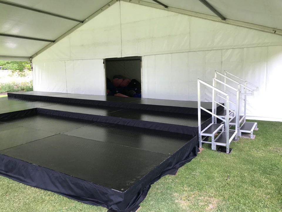 Black skirted stage with handrail steps inside an open marquee on grass